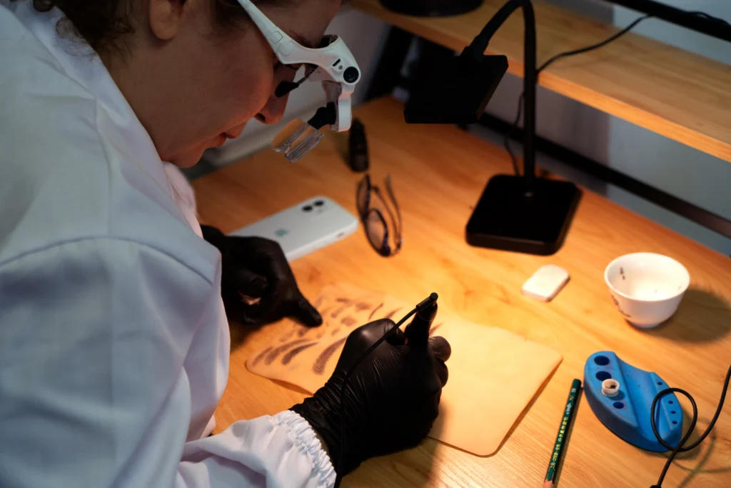 Close-up of a PMU artist practicing nano brows on synthetic skin during a training session. The artist wears magnifying loupes and black gloves while using a tattoo machine to create realistic hair strokes. The workspace includes a pigment cup, pencil, and desk lamp, typical of a Hyper Real Academy course. Ideal for learning powder brows, lip blush, SMP, or eyeliner techniques using RealTint® pigments.