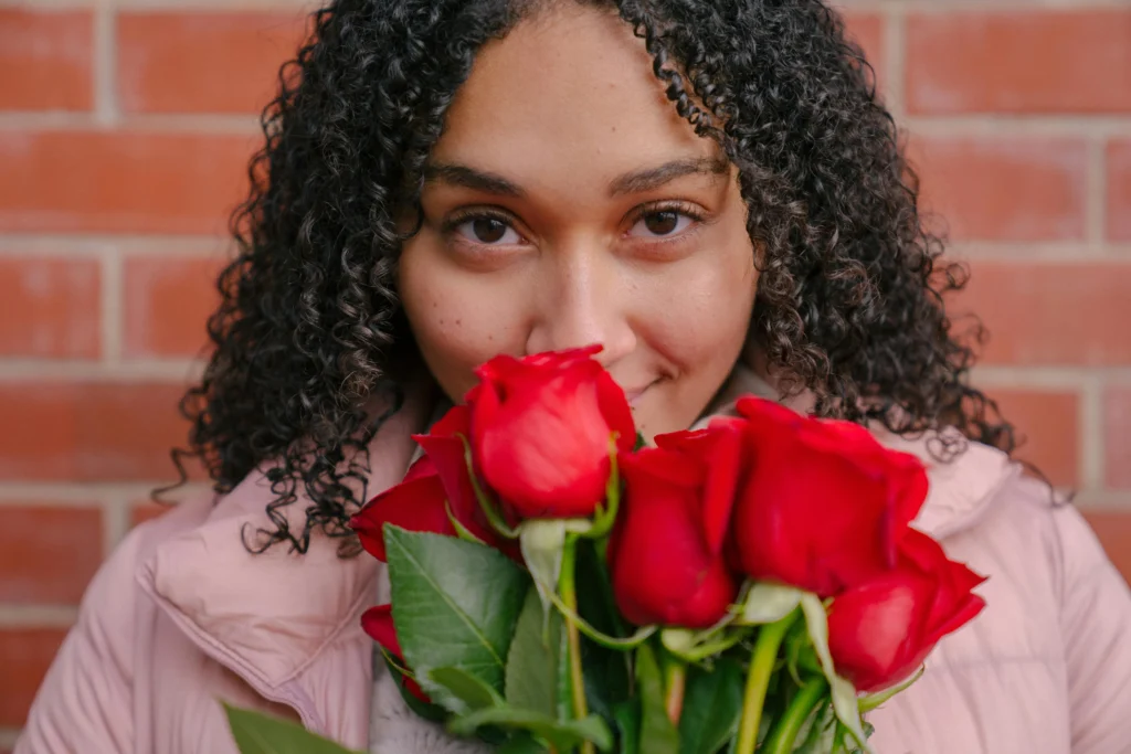 Woman with curly hair holding red roses against brick wall, beauty model for Hyper Real Academy PMU training.