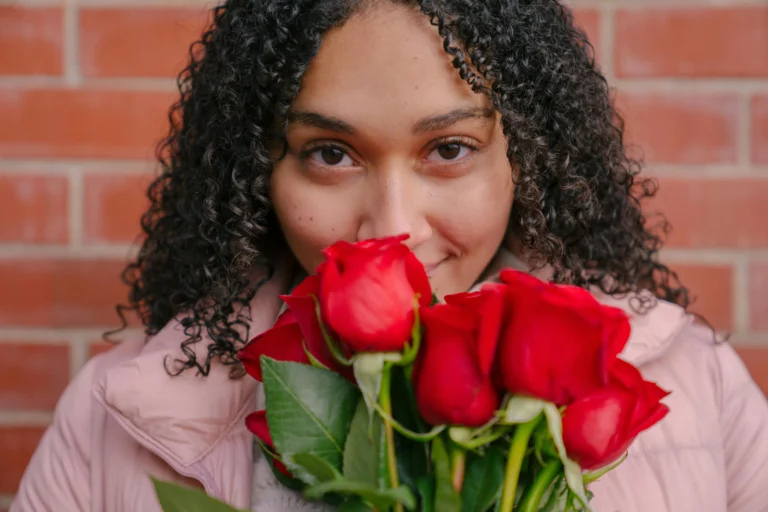 Woman with curly hair holding red roses against brick wall, beauty model for Hyper Real Academy PMU training.