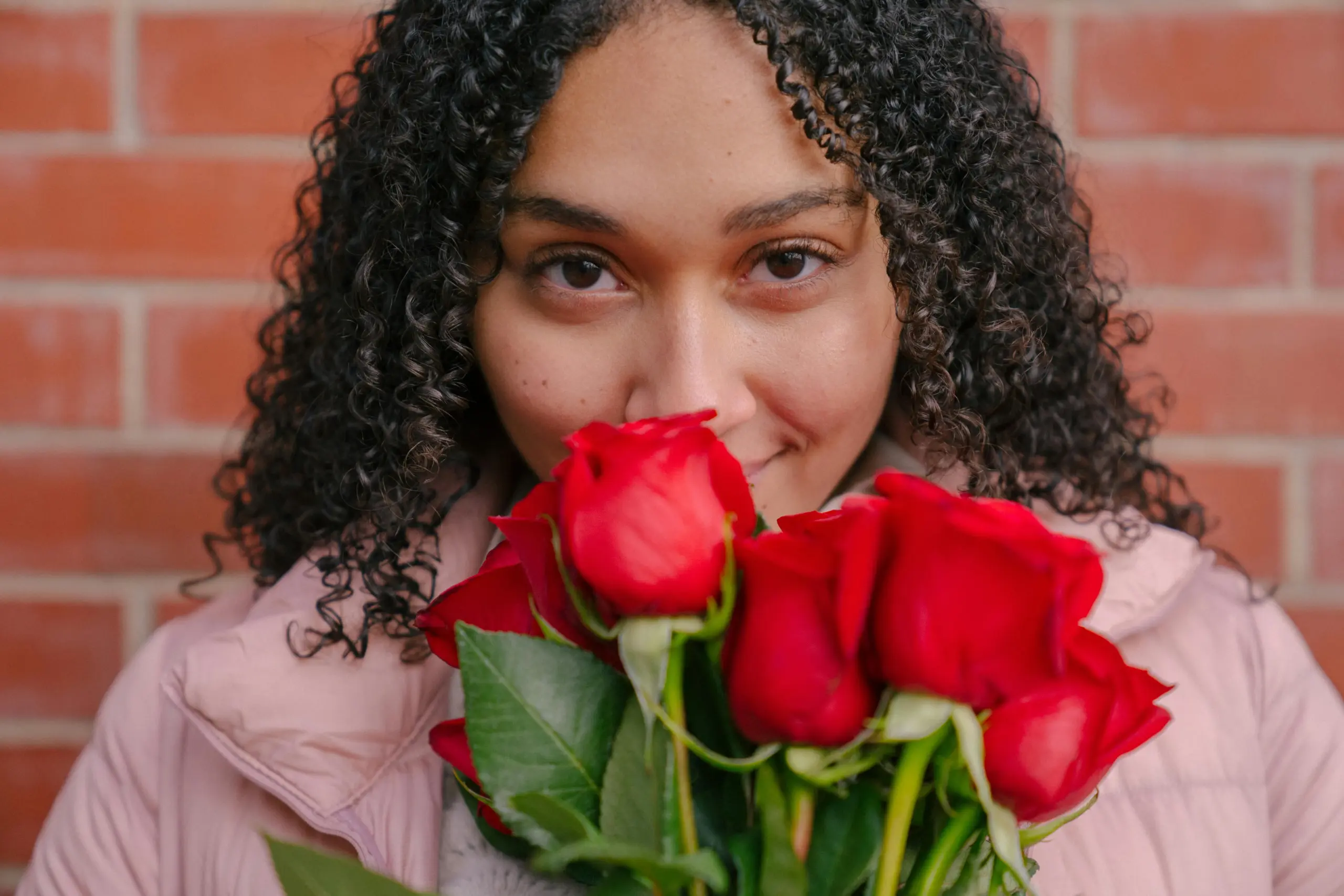 Woman with curly hair holding red roses against brick wall, beauty model for Hyper Real Academy PMU training.