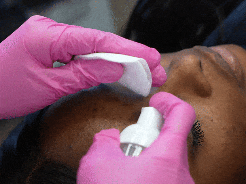PMU training demo: Pink gloved hands spray antimicrobial foam on cotton pad to prep skin for nano brows or powder brows.