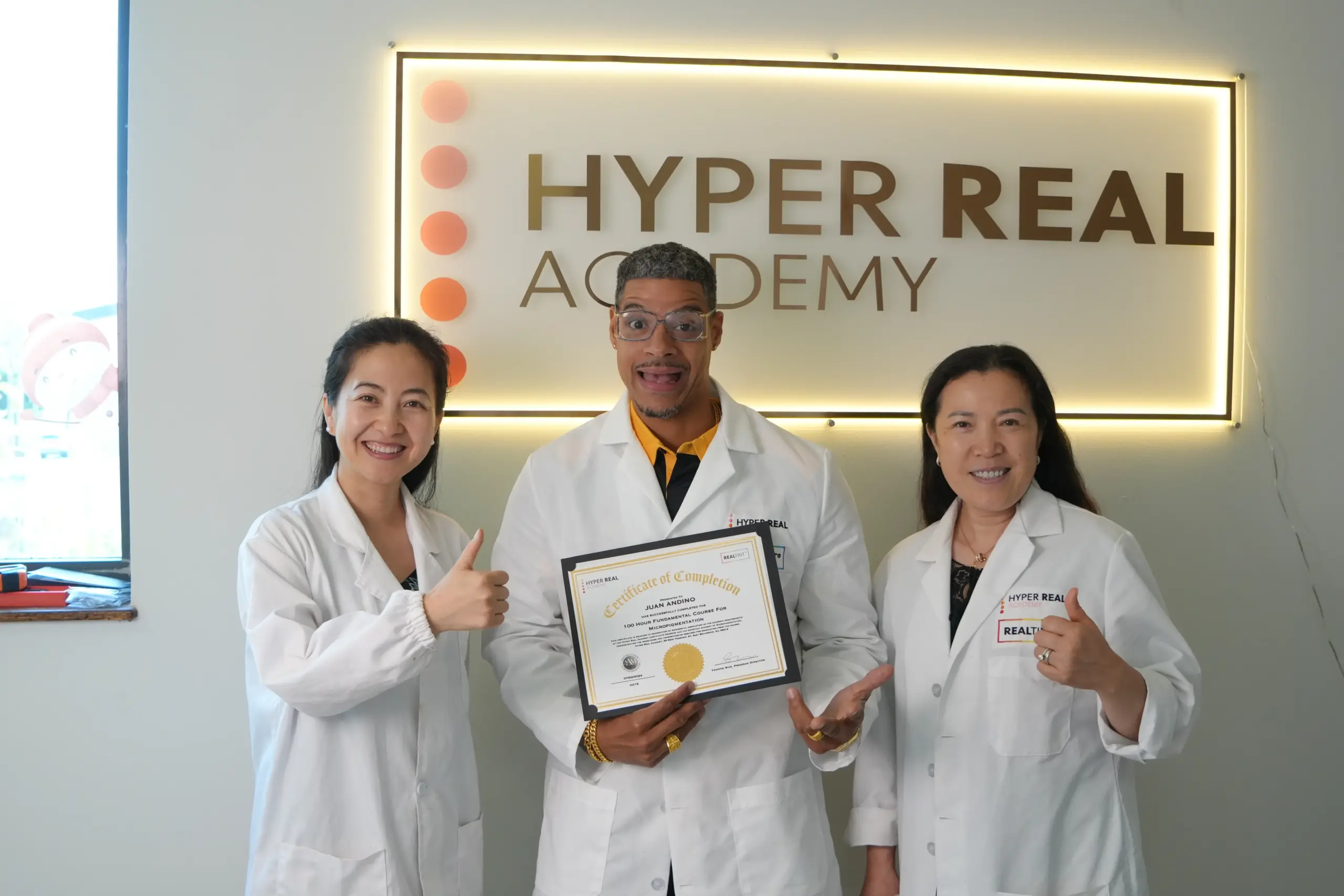 Hyper Real Academy PMU training graduation. Student holds micropigmentation certificate with instructors. RealTint.