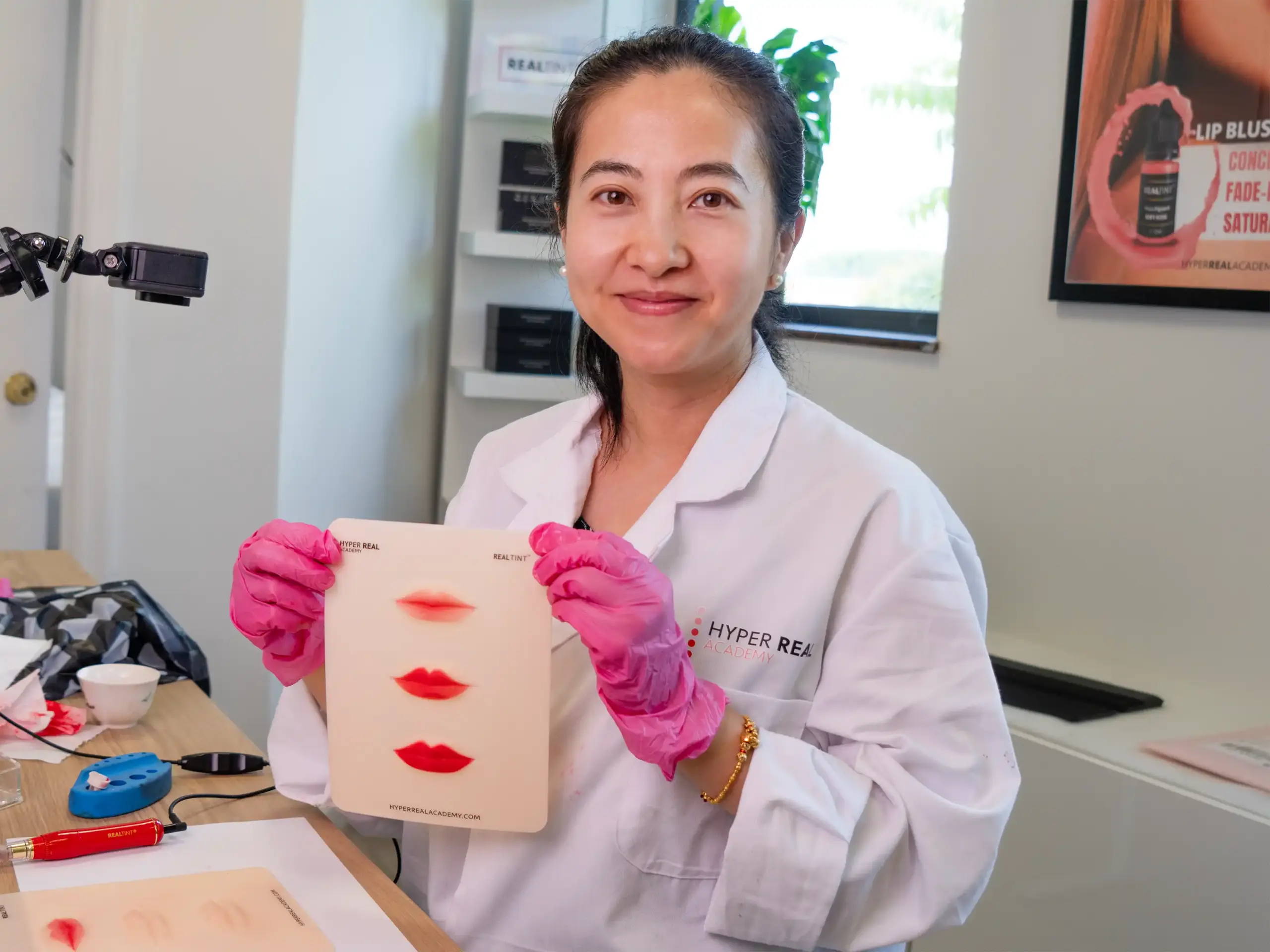 Silicone practice skin with lip blush shape and faint hair strokes. Red tattoo machine pen, ink bottle, and pink gloves visible. Nano brows or machine hair strokes training setup, not traditional microblading.