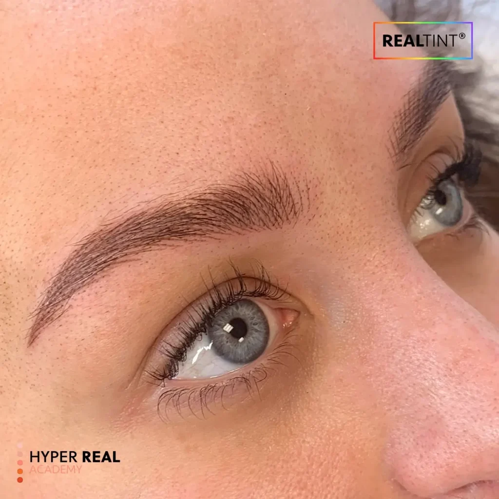 During nano brows procedure: technician's gloved hands using rotary pen to create individual hair strokes on client's eyebrow. Fresh dark pigment visible, red gauze wiping excess. Active pigment implantation stage.