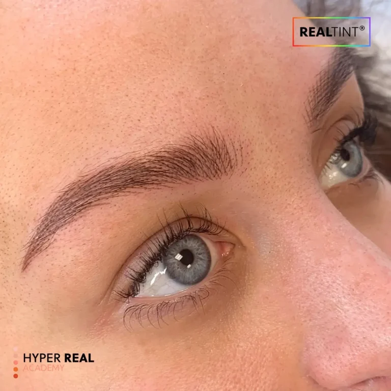 During nano brows procedure: technician's gloved hands using rotary pen to create individual hair strokes on client's eyebrow. Fresh dark pigment visible, red gauze wiping excess. Active pigment implantation stage.