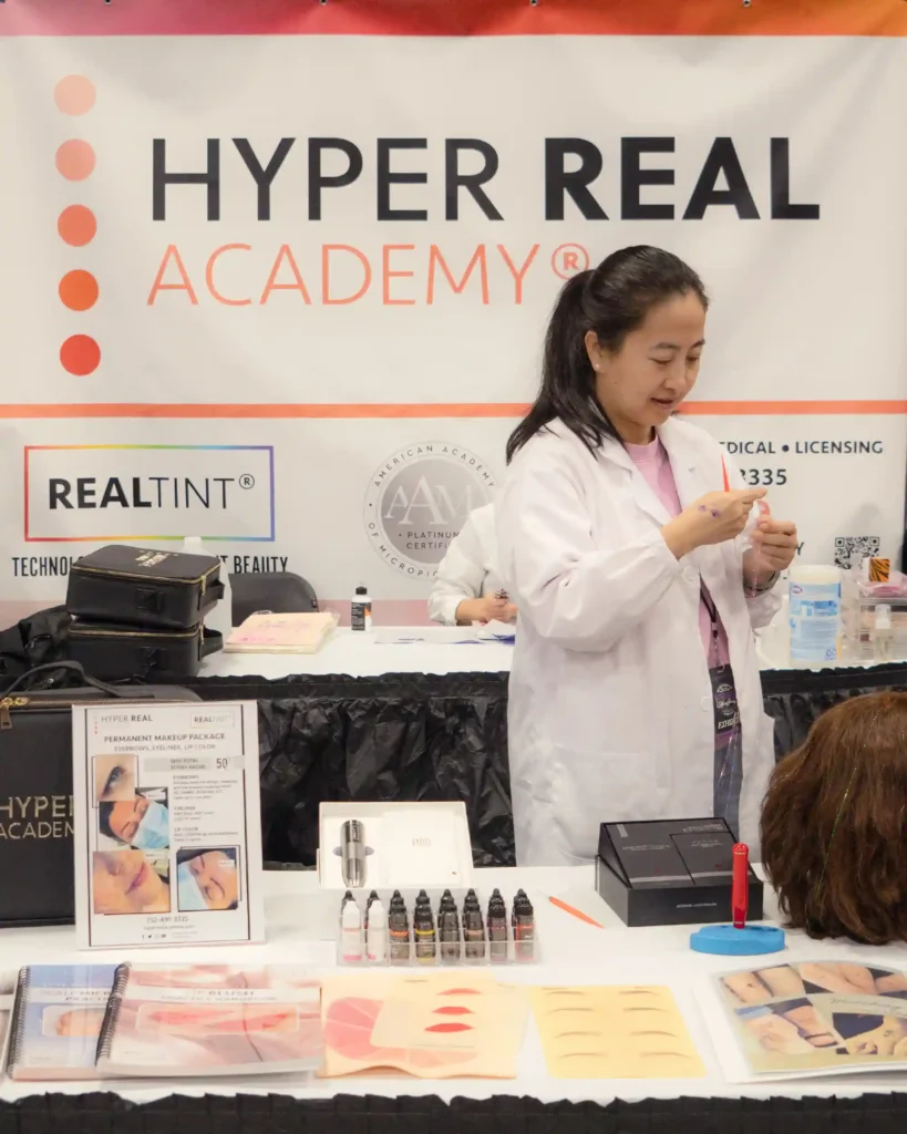 PMU procedure in progress at trade show or competition. Technician in white coat, pink mask performing microblading or nano brows on client. Black curtain background, 'TATTOO REAL' watermark. Shows PMU competition or workshop demonstration.