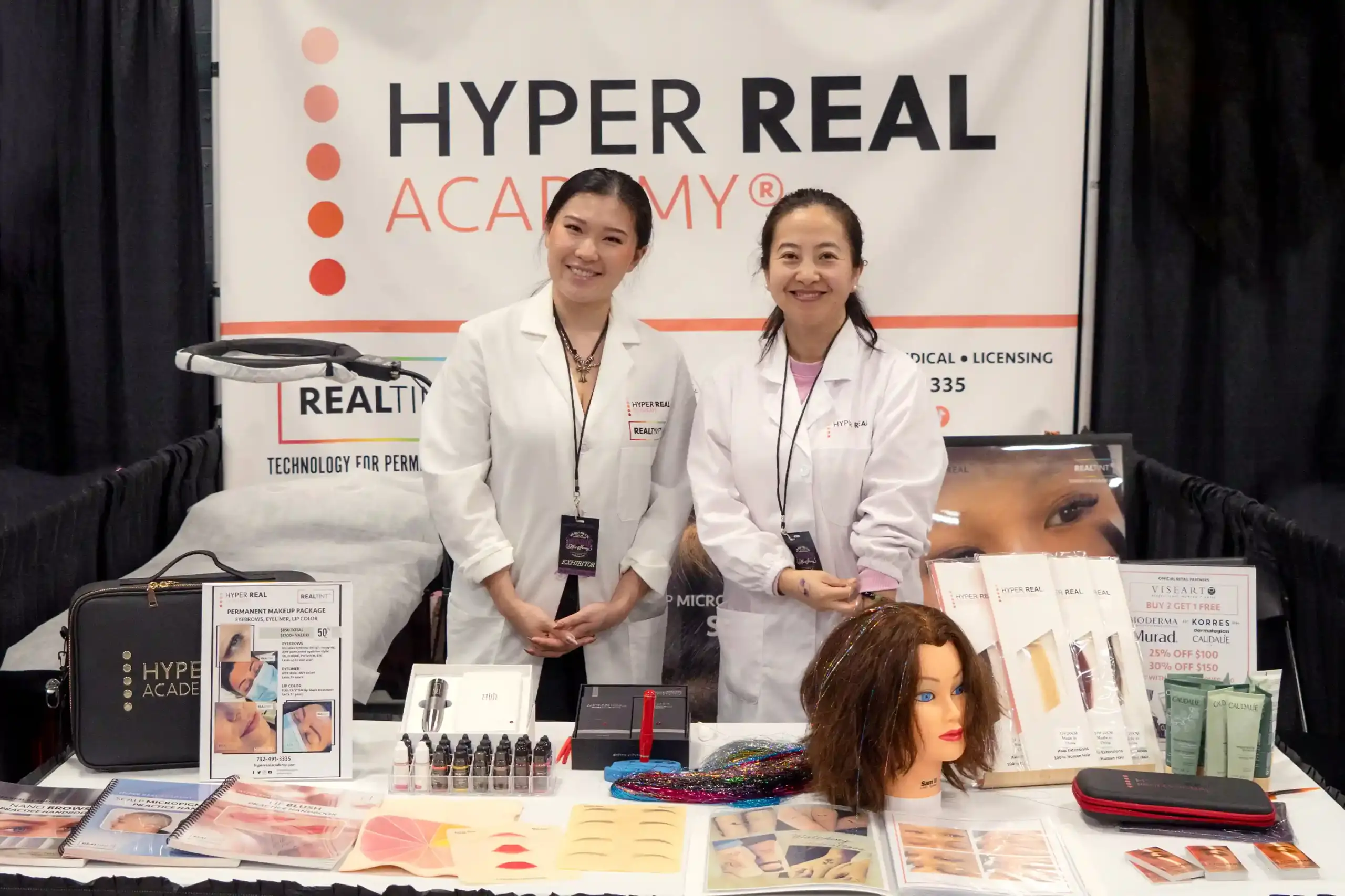 Trade show booth for Hyper Real Academy. Woman in white coat demonstrating products at display table with Real Tint branding, brochures, and equipment. Beauty expo or educational convention promotional photo.