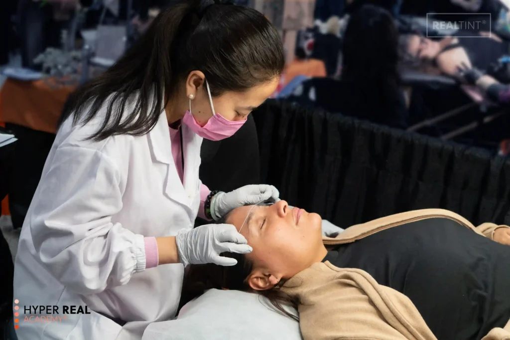 PMU procedure in progress. Technician in white gloves performing microblading or nano brows on client's eyebrow. Client lying down with eyes closed. Hyper Real branding. Shows active PMU eyebrow enhancement procedure.