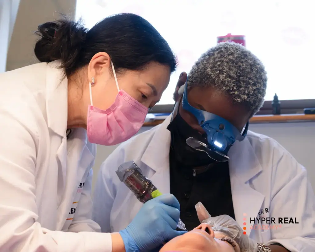 PMU training session. Three women in white lab coats and pink masks working on client's eyebrow. One operating PMU machine, two observing closely. Student photo or instructional demonstration with 'HYPER REAL' watermark.