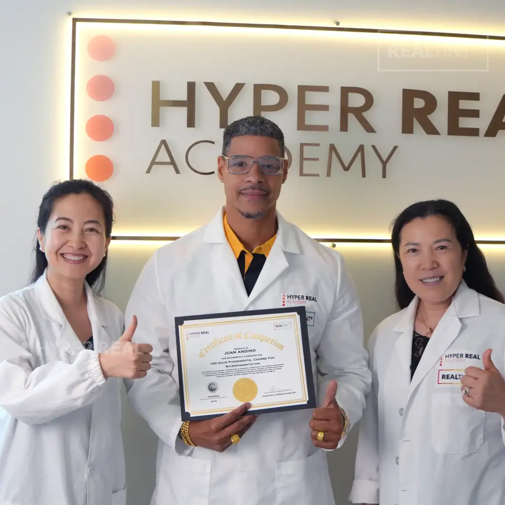 Hyper Real Academy instructor performing brow mapping for a PMU procedure. A professional in a white coat and pink mask uses a pencil to design eyebrows on a client, demonstrating techniques for nano brows, powder brows, and permanent makeup. RealTint® branding visible.