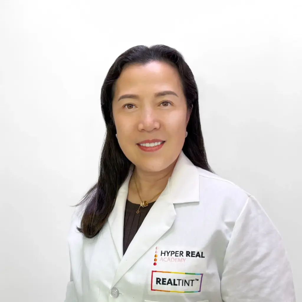 Professional headshot of woman in white lab coat with 'HYPER REAL REALITY' name tag. Medium-shot portrait against white background. Promotional photo for PMU practitioner or academy staff member.
