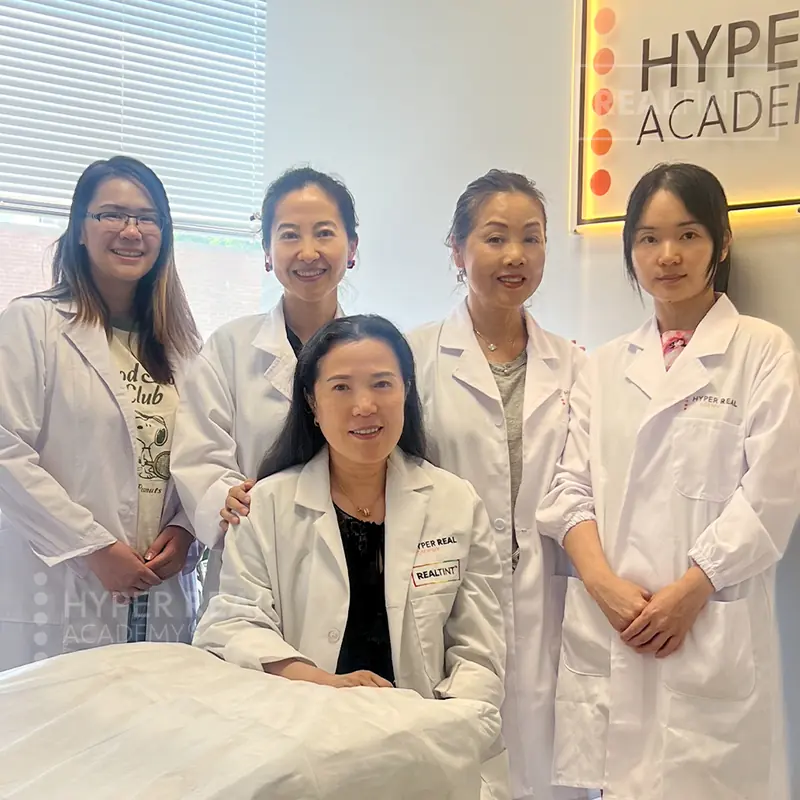 Hyper Real Academy graduation - student in center holding framed certificate with gold seal, flanked by two instructors in white coats giving thumbs up. 'HYPER REAL ACADEMY' and 'HYPER REAL BEAUTY' signage visible.