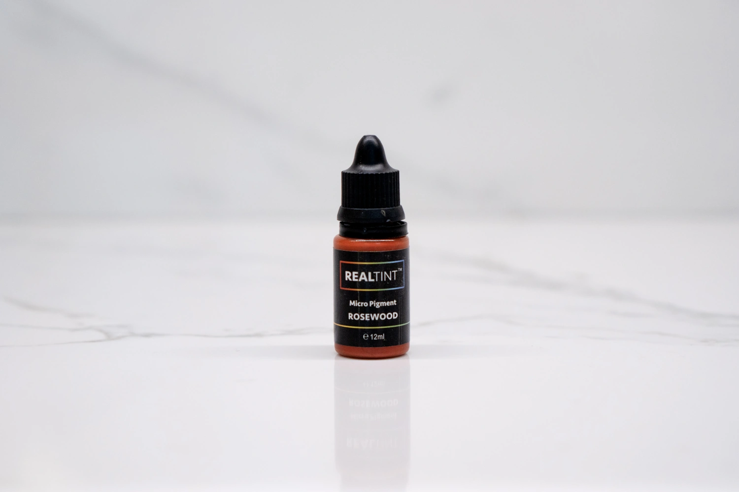RealTint(R) Rosewood micro pigment, a 12 ml dropper bottle of warm reddish-brown permanent makeup pigment on a white marble surface